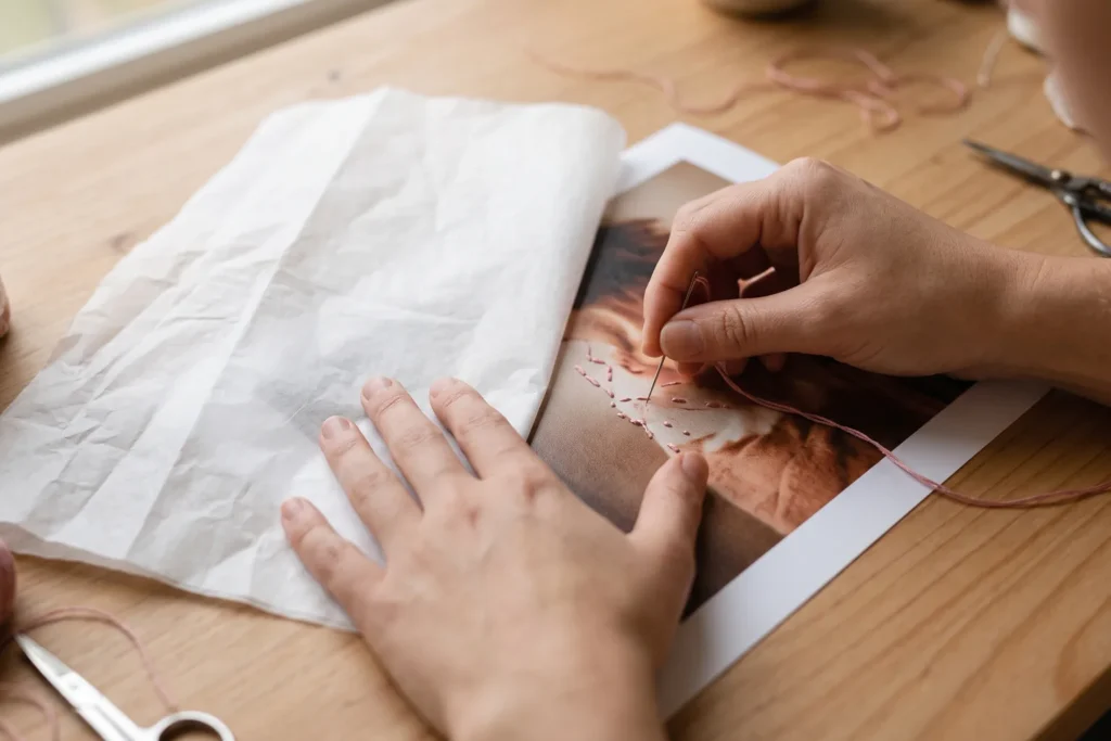 Mano apoyada sobre un papel de seda colocado encima de una fotografía mientras borda con la otra mano